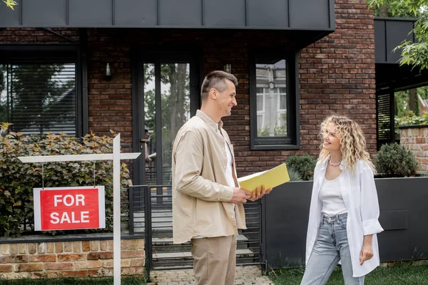 Alegre propiedad inmobiliaria con carpeta hablando con la mujer cerca de casa moderna en la ciudad - foto de stock