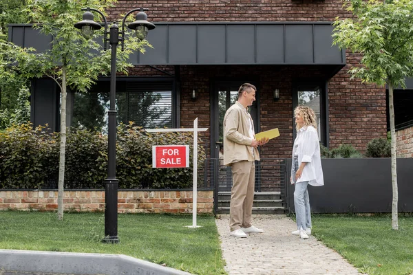 Smiling woman talking to real estate agent near modern cottage and signboard with for sale lettering — Stock Photo