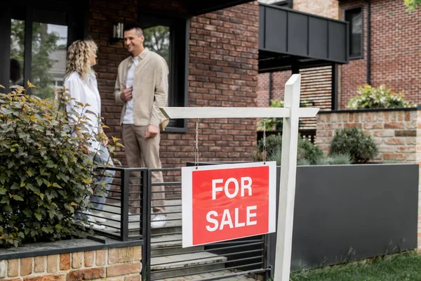 Gioioso e di successo broker immobiliare parlando con la donna vicino casa contemporanea in vendita sulla strada — Foto stock