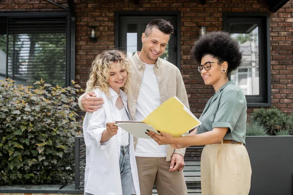African american property realtor showing documents to happy couple near new modern cottage — Stock Photo