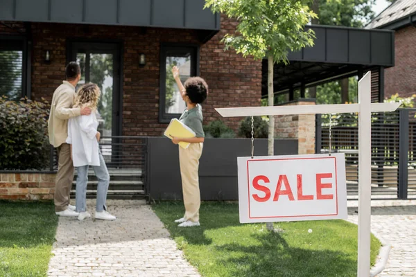 African american real estate agent showing house to hugging couple near for sale signboard — Stock Photo