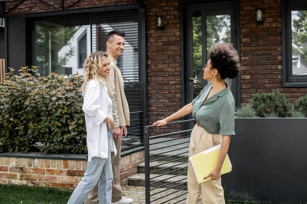 Joyful couple talking to african american property realtor near modern building on urban street — Stock Photo