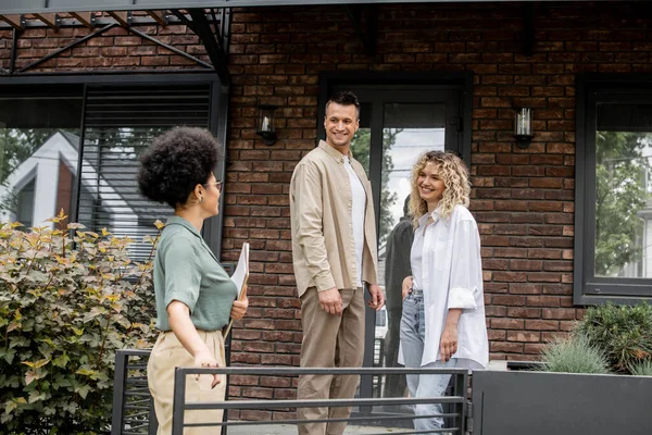 Afrikanisch-amerikanischer Immobilienmakler im Gespräch mit lächelndem Paar in der Nähe eines neuen modernen Häuschens zum Verkauf — Stockfoto