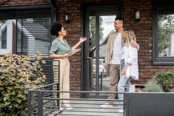 Positiv afrikanisch-amerikanischer Immobilienmakler mit Folder, der glücklichen Eigentümern zeitgenössisches Ferienhaus zeigt — Stockfoto