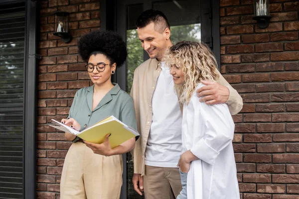 Agente inmobiliario afroamericano mostrando documentos a los compradores encantadores abrazos cerca de nueva casa - foto de stock