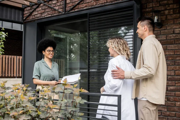 Cheerful african american property realtor with folder talking to clients near new cottage — Stock Photo