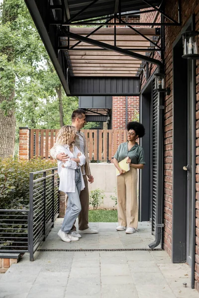 Sorridente agente immobiliare africano americano con cartella che parla con coppia di nuovi proprietari vicino casa — Foto stock