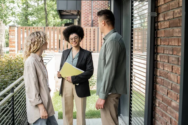 Agente inmobiliario afroamericano positivo con documentos que hablan con un par de clientes cerca de casa de campo - foto de stock