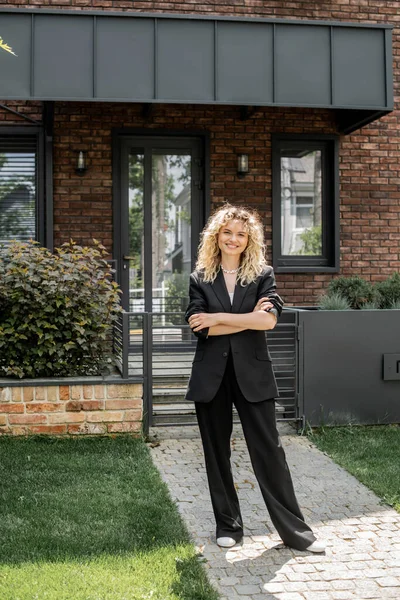 Fiducioso e attraente agente immobiliare in piedi con le braccia piegate vicino casa moderna sulla strada urbana — Foto stock