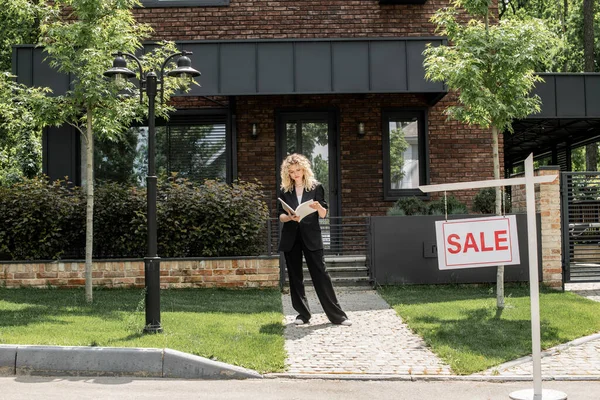 Intera lunghezza di elegante agente immobiliare in piedi con cartella vicino alla casa urbana contemporanea in vendita — Foto stock
