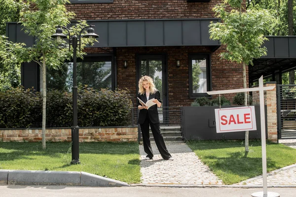 Full length of stylish real estate agent standing with folder near contemporary urban house for sale — Stock Photo