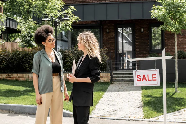 Блондинка агент по недвижимости разговаривает с африканским американским клиентом рядом с домом и на продажу вывески — стоковое фото