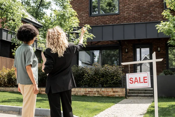 Rückansicht des blonden Maklers, der mit der Hand zeigt, zeigt Ferienhaus zum Verkauf an afrikanisch-amerikanische Frau — Stockfoto
