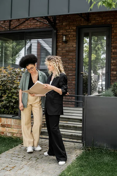 Agente imobiliário loira mostrando documentos para cliente afro-americano perto de casa moderna para venda — Fotografia de Stock