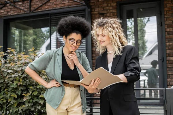 Property agent showing documents to african american client standing with hand on hip near new house — Stock Photo