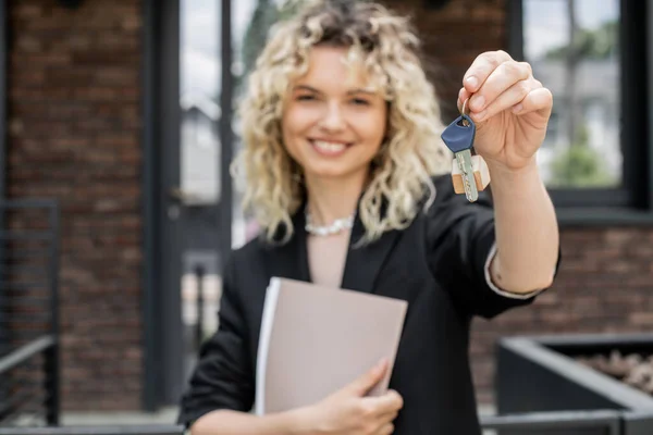 Blurred property agent smiling and looking at camera while holding folder and key from new house — Stock Photo