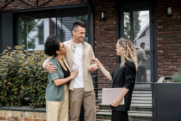 Successful property agent giving key to happy multiethnic couple near house on city street — Stock Photo