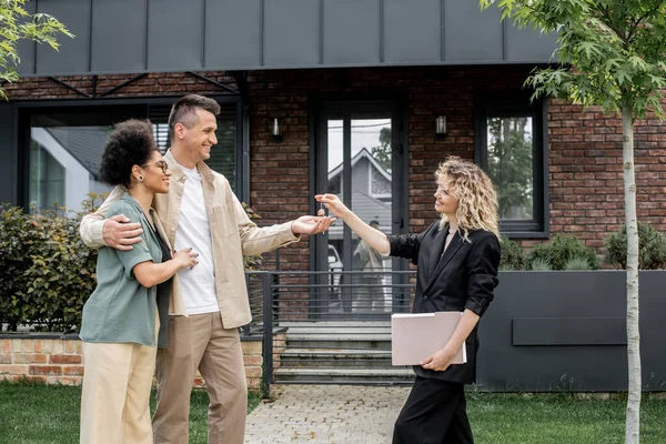 Real estate broker with folder giving key to overjoyed interracial couple near new city cottage — Stock Photo