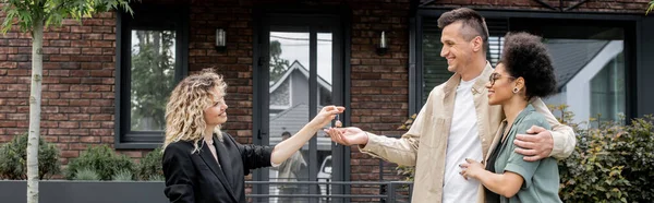 Glückliches gemischtrassiges Paar nimmt Schlüssel aus neuem zeitgenössischen Ferienhaus von Immobilienmakler, Banner — Stockfoto