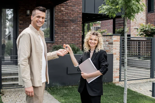 Blonde Immobilienmaklerin hält Ordner in der Hand und übergibt Schlüssel von neuem Ferienhaus an freudige Klientin — Stockfoto