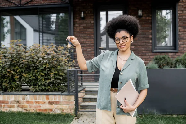 Agent immobilier afro-américain avec clé et dossier souriant à la caméra près du nouveau chalet — Photo de stock