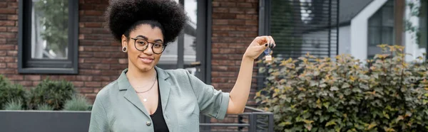 Sorridente agente immobiliare africano americano in occhiali che mostra la chiave della nuova casa all'aperto, banner — Foto stock