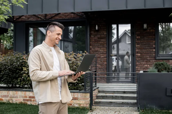 Gioioso broker immobiliare utilizzando laptop vicino cottage moderno sulla strada urbana — Foto stock