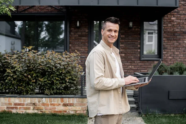 Agente immobiliare di successo con laptop sorridente alla fotocamera vicino edificio sulla strada urbana — Foto stock
