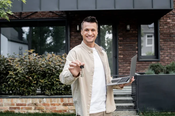 Agente imobiliário alegre com laptop mostrando a chave da nova casa enquanto está ao ar livre — Fotografia de Stock