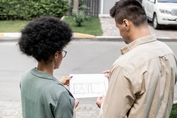 Back view of multicultural real estate agents looking at blueprint of building while outdoors — Stock Photo