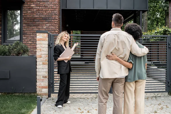 Lächelnder blonder Immobilienmakler mit Folder, der dem multiethnischen Paar das neue Stadthaus zeigt — Stockfoto