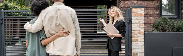 Agent immobilier blonde positive avec dossier pointant vers une nouvelle maison près d'un couple multiculturel, bannière — Photo de stock