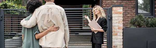 Positiver blonder Makler mit Ordner, der auf neues Haus in der Nähe multikultureller Paare zeigt, Banner — Stockfoto