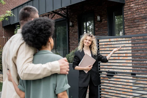 Fröhlicher Immobilienmakler mit Folder, der multiethnischen Käuferpaaren das neue Stadthaus zeigt — Stockfoto