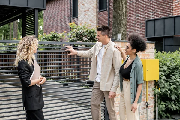 Задоволена багатоетнічна пара, що стоїть біля агента з нерухомості і паркану нового міського котеджу — стокове фото