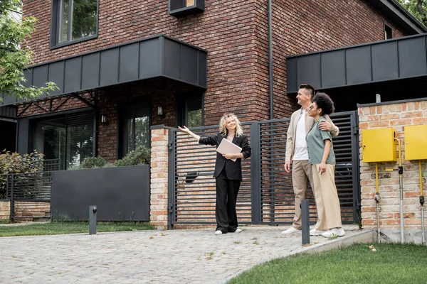Immobilienmakler zeigt mit der Hand, während er dem multiethnischen Paar sein neues Ferienhaus zeigt — Stockfoto