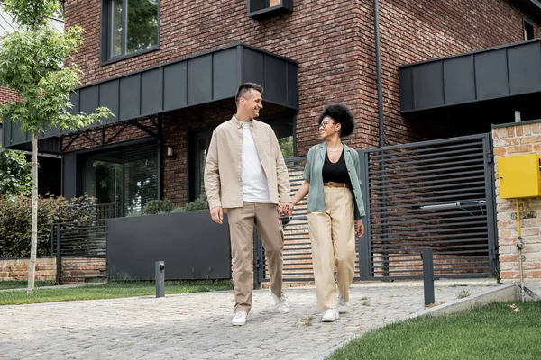 Happy interracial owners of new cottage holding hands and looking at each on city street, banner — Stock Photo