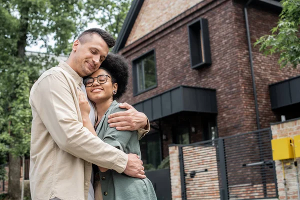 Freudiges multiethnisches Paar, das sich vor dem neuen eigenen Häuschen an der städtischen Straße umarmt, Banner — Stockfoto