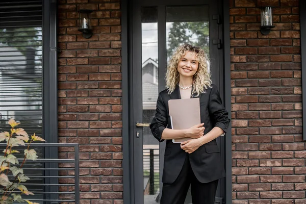 Blonde und attraktive Immobilienmaklerin hält Ordner in der Hand und lächelt in die Kamera in der Nähe des Stadthauses — Stockfoto
