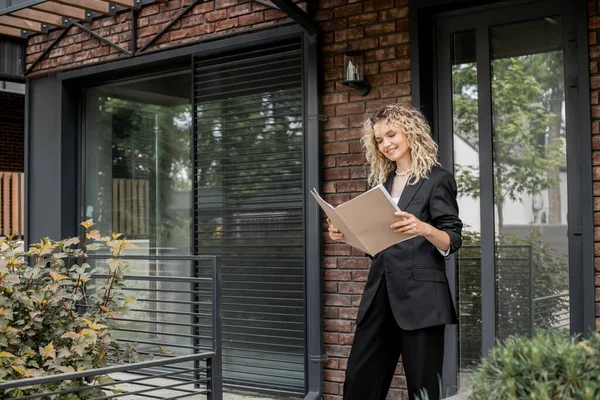 Lächelnder blonder Immobilienmakler beim Betrachten von Dokumenten in Ordnern in der Nähe eines modernen neuen Hauses — Stockfoto