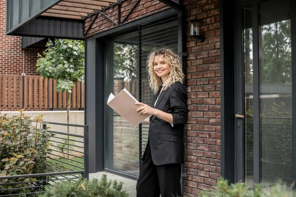 Joyful blonde real estate broker with folder looking at camera near modern city house — Stock Photo