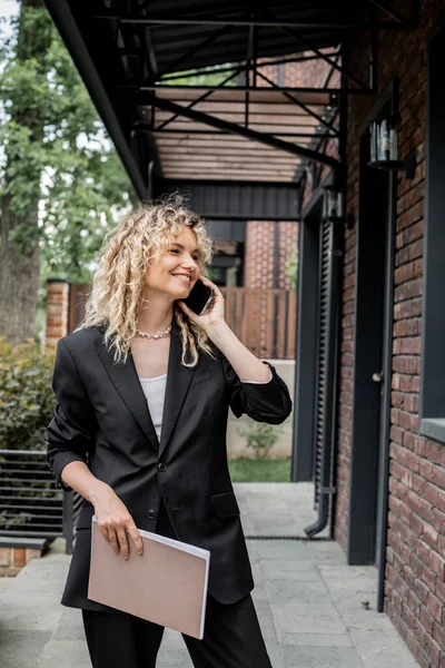 Pleased and attractive real estate agent holding folder and talking on smartphone near new cottage — Stock Photo