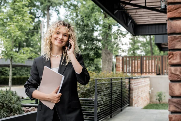Positive real estate agent with folder talking on smartphone and looking at camera near house — Stock Photo