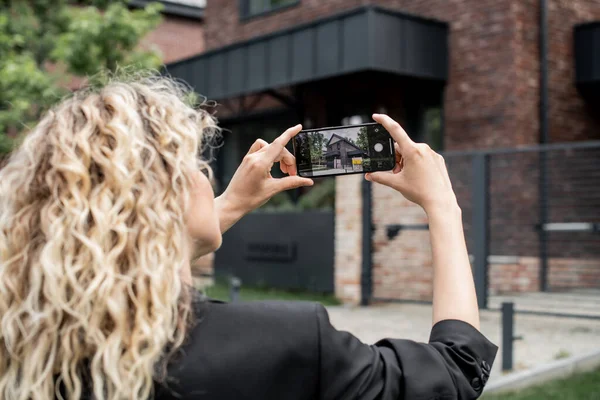 Blonde real estate broker with mobile phone taking photo of contemporary cottage on city street — Stock Photo