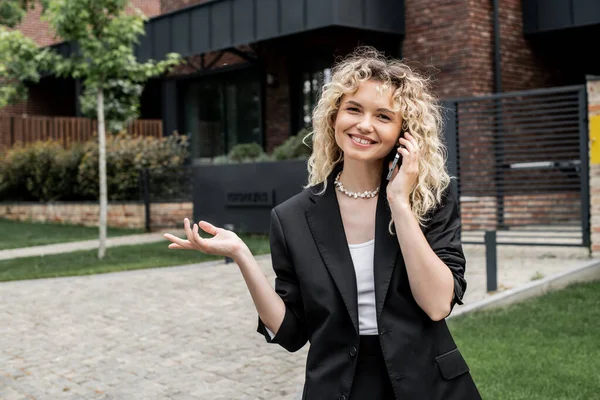 Blonde Immobilienmaklerin zeigt mit der Hand und lächelt in die Kamera, während sie auf dem Smartphone spricht — Stockfoto