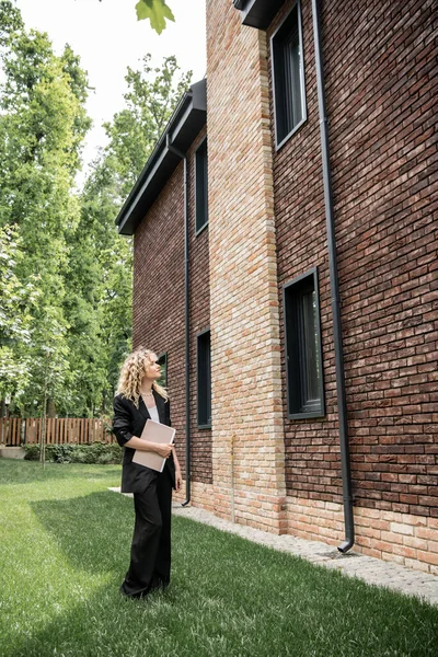 Full length of blonde real estate broker with folder looking at new cottage on city street — Stock Photo
