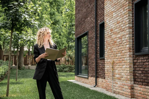 Blonde and stylish real estate agent holding folder with documents and looking at city cottage — Stock Photo