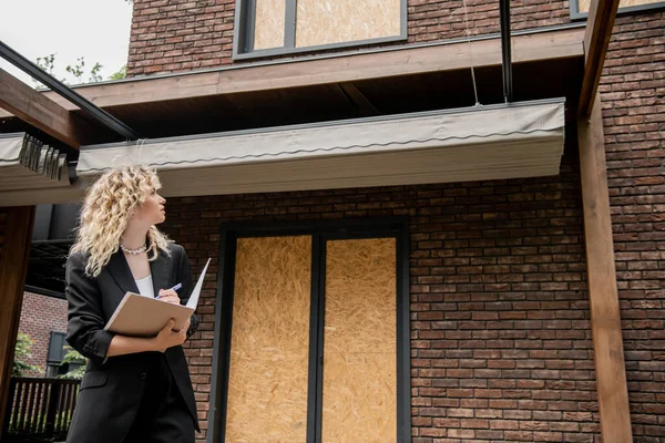 Блондинка агент по недвижимости глядя на новое здание и писать в папке на открытом воздухе — стоковое фото