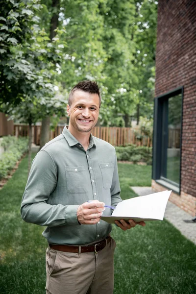 Überglücklicher Immobilienmakler blickt in die Kamera, während er mit Aktenordner und Stift in der Nähe von Ferienhaus steht — Stockfoto