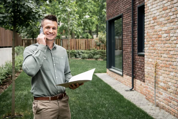 Cheerful real estate broker with folder talking on smartphone and looking at camera near house — Stock Photo